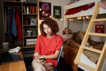 Portrait of curly haired young woman using smartphone in dorm room and scrolling social media, copy...