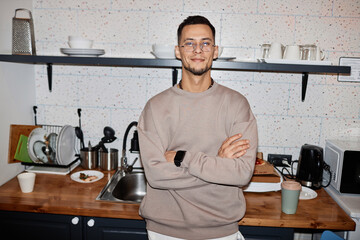 Waist up portrait of confident young man wearing eyeglasses standing with arms crossed in college dorm kitchen, flash