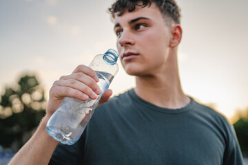 caucasian man teenager open plastic bottle of water outdoor in sunny day drink while hold basketball