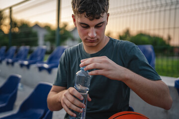 caucasian man teenager open plastic bottle of water outdoor in sunny day drink while hold basketball