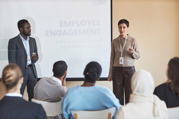 Confident mature female HR manager standing by whiteboard in front of audience and explaining one of presentation points