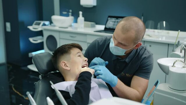 Young patient with toothache opening mouth wide, showing dental problem, professional dentist in medical mask and gloves examining oral cavity of boy. High quality 4k footage