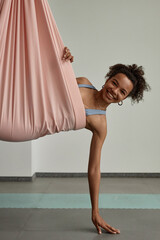 Vertical portrait of Black young woman enjoying aerial yoga in pink hammock and smiling at camera