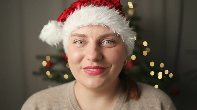 Happy smiling Caucasian beautiful woman in Santa hat looking at camera, smiling and chatting via online video call beside Christmas tree at home. High quality FullHD footage