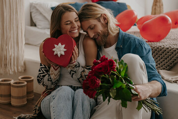 Loving couple embracing and holding roses and gift while leaning on bed with heart shape balloons 