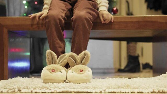 Cute boy grabbing his new bunny slippers and putting them on on his feet. Jumping from excitement in front of Christmas tree. High quality 4k footage