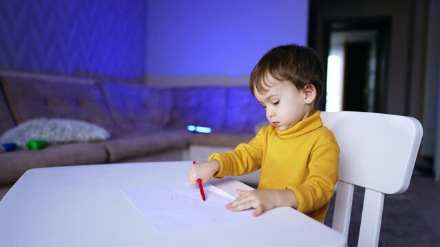 Charming little baby sitting at the desk drawing. Adorable child is focused on the hobby. Blurred backdrop.
