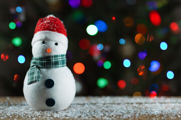 Snowman and Christmas tree. Winter home decorations. Holiday traditions and gifts. Merry Christmas. Snowman toy. Christmas tree lights bokeh on background. Fake snow on wooden desk. Happy New Year
