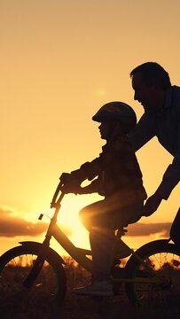 Father teaches child wearing safety helmet to ride bicycle Family in park. Child rides bicycle. Father helps his daughter ride bike. Kid, dad play together, sunset. Child dream learns to ride bicycle