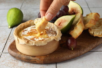 Woman dipping bread into baked brie cheese at light tiled table, closeup