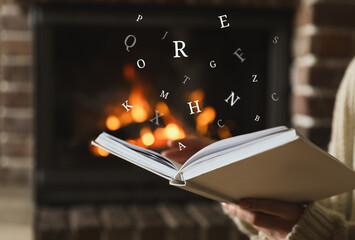 Woman reading book with letters flying over it near fireplace at home, closeup