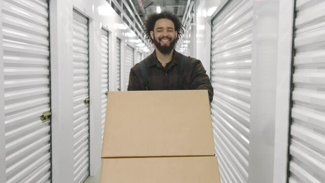 Happy male worker, dark-skinned man transporting large carton boxes, leisurely walking along long white illuminated corridor, narrow passage, shutter doors. High quality 4k footage