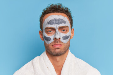Young serious man portrait with clay facial mask posing isolated on blue backdrop, spa treatments concept, copy space