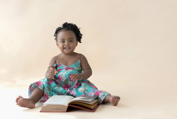 A 2-year-old Nigerian baby girl with beautiful curly hair, sitting on the floor, holding a yellow...