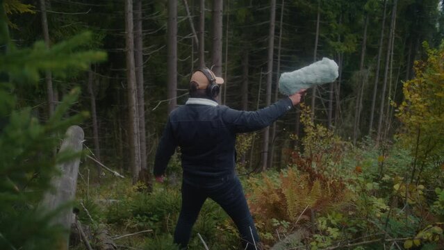 Sound engineer wearing headphones records sounds of nature for movie in the forest using professional audio equipment. Caucasian man works with furry windscreen microphone outdoors. Film production.