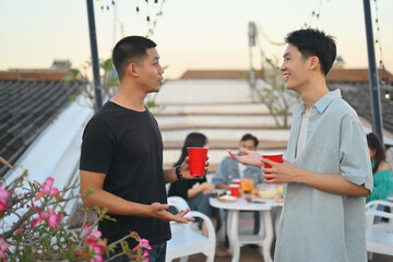 Two young handsome men having drinks and chatting at the rooftop party in the evening.