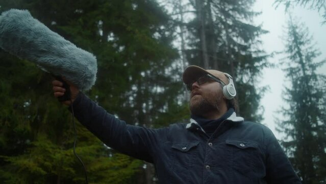 Caucasian man wearing headphones records sounds of nature for movie in coniferous wood using furry windscreen microphone. Sound engineer works with professional audio equipment outdoors. Slow motion.