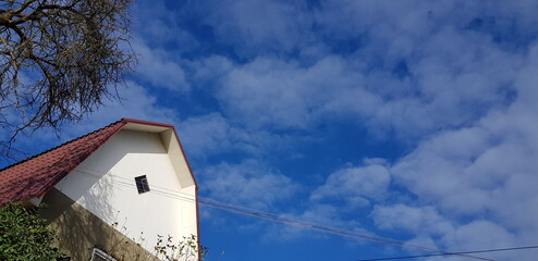 Panorama of blue sky in white and fluffy clouds over a white house with a red roof and bare treetop (bottom, left) in the sun in winter (winter landscape, sky texture, house in the sky).