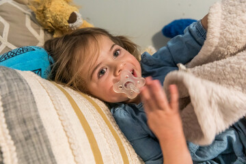 Toddler girl sucking on pacifier in bed with blanket
