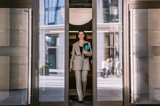poised young woman exits a modern building, dressed in a tailored beige suit, carrying a blue notebook