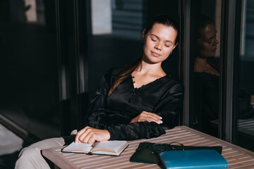 Peaceful woman in a black blouse sits with closed eyes, sunlight caressing her face, with a book on...
