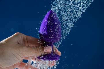 Woman washing purple anal plug under shower on blue background. 