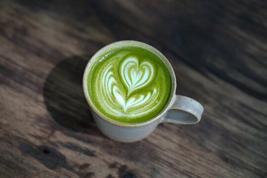 A cup of green matcha latte with heart shaped latte art on wooden table.