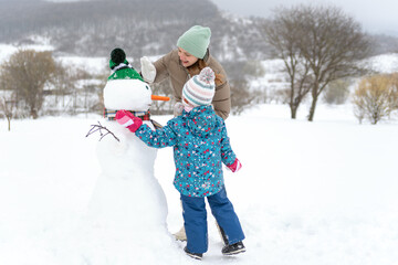 Fototapeta na wymiar Mother and Toddler build large snowman with carrot nose. Kid during stroll in a snowy winter park.