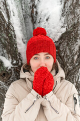 Young blue eyes woman warms up hands in woolen hat and mittens, cold weather. Under snow-covered tree, winter park