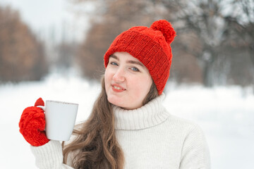 Smiling young woman in red knitted hat and mittens in winter park. Happy girl with cup in hands on...