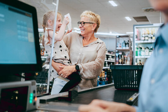 Happy senior woman carrying cheerful granddaughter holding bill at supermarket