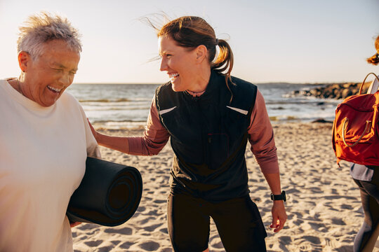 Happy female coach laughing with senior woman holding exercise mat at beach