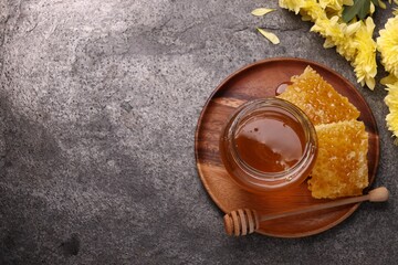 Obraz na płótnie Canvas Sweet golden honey in jar, dipper, pieces of honeycomb and chrysanthemum flowers on grey textured table, flat lay. Space for text