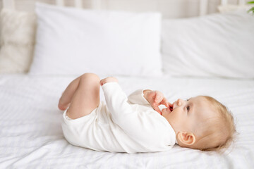 smiling baby 6 months old blond boy lies on a large bed in a bright bedroom and plays with his legs...