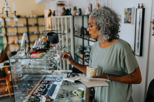 Senior female entrepreneur with diary and coffee cup examining jewelry at antique shop