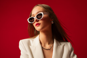 Fashionable confident woman wearing trendy white sunglasses, suit blazer, posing on red backdrop....