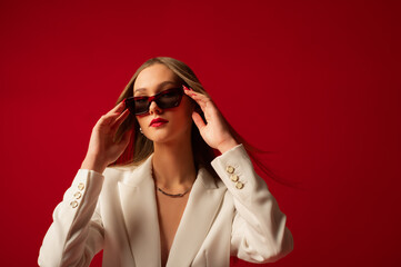 Fashionable confident woman wearing trendy sunglasses, white suit blazer, posing on red backdrop....