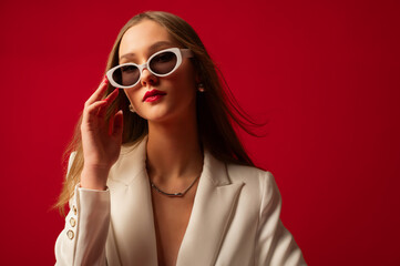 Fashionable confident woman wearing trendy white sunglasses, suit blazer, posing on red backdrop. Studio fashion portrait. Copy, empty space for text