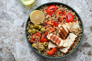 Plate of quinoa with grilled haloumi, bell pepper and green olives, horizontal shot on a light-brown granite background, high angle view