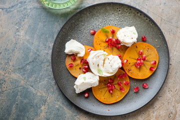 Plate with fresh persimmon slices, torn mozzarella and pomegranate seeds, high angle view on a beige and grey granite background, horizontal shot