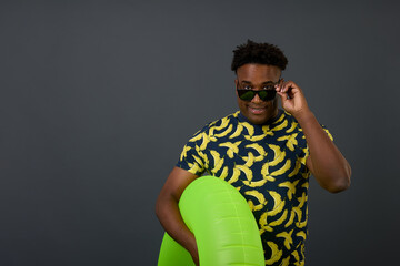 Satisfied dark-skinned guy, a tourist, resting on a gray background, adjusts sunglasses with an inflatable circle in his hand. African young handsome man posing in fashionable t-shirt