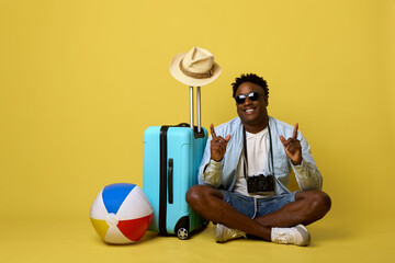 Positive stylish African in fashionable sunglasses is sitting next to a suitcase and leisure paraphernalia on a yellow background. A young cheerful guy is photographed before a summer trip - obrazy, fototapety, plakaty