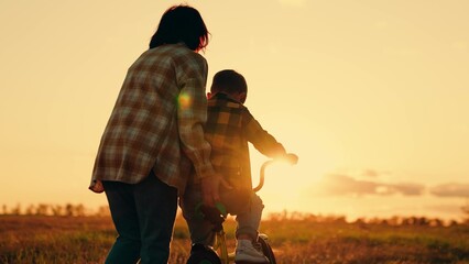 Mom teaches her son to ride bike in park at sunset. Family day out in nature. Mother little son learn to ride bike together in summer. Boy, his mother learn to ride bicycle in meadow in sun. Teamwork
