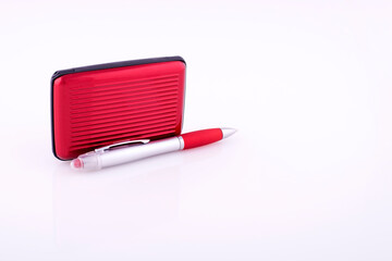 Red plastic credit card case and ballpoint pen on white reflective background.