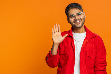 Young Indian man smiling friendly at camera, waving hands gesturing hello greeting, goodbye welcoming with invitation hospitable expression. Arabian Hindu guy isolated on orange background. Copy-space - obrazy, fototapety, plakaty