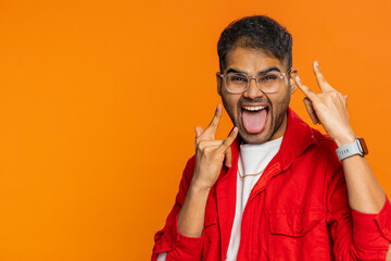 Overjoyed happy excited Indian man showing rock n roll gesture by hands, cool sign, shouting yeah with crazy expression, dancing, rejoicing victory win success. Guy on orange background. Copy-space