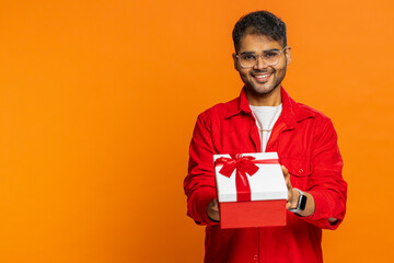 Smiling Indian man presenting birthday gift box stretches out hands, offer wrapped present career bonus celebrating party promotion discount sale. Arabian guy isolated on orange background. Copy-space