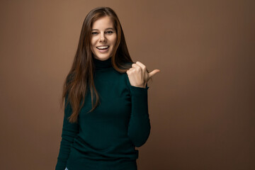 Confident young Caucasian woman with long brown hair points with her hand towards an advertisement on a brown background