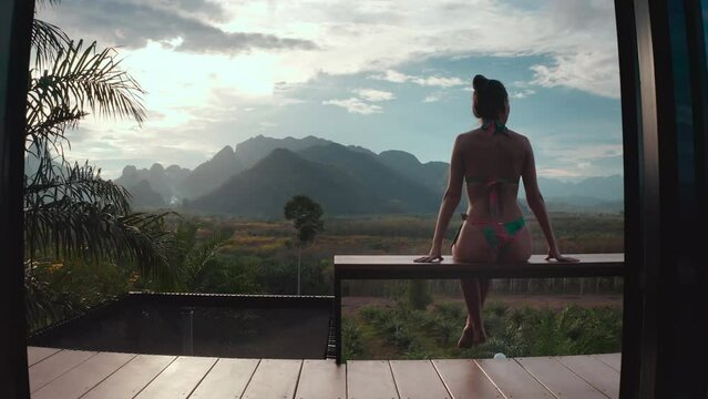 Woman relax in holiday hut villa with mountain wild nature view. Female in bikini enjoy tropical landscape sitting on cottage balcony. Outdoor lifestyle travel on summer vacation. Back view, zoom in