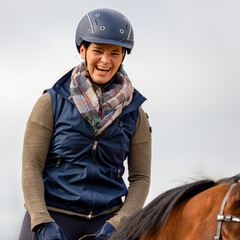 Rider woman on the horse with a riding helmet looks happily laughing into the camera.